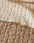 A close-up of a bed with two quilts: on top is Coyuchi Inc’s Blossom Hand Stitched Quilt in light beige with a grid pattern, while the bottom quilt shows a floral design with orange and green accents.