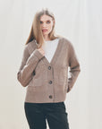 A woman with straight, light brown hair wears the Jumper 1 2 3 4/CR2 Guernsey Cardigan in fawn grey over a white shirt and dark pants, standing against a light, neutral background.