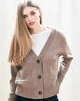 A woman with straight, long blonde hair wears a fawn grey Guernsey Cardigan by Jumper 1 2 3 4/CR2 over a white shirt, standing against a light background and looking at the camera with a neutral expression.