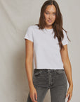 A woman with long brown hair wears a plain white Perfectwhitetee Springsteen Baby Tee and high-waisted black jeans, standing against a neutral light background.
