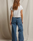 A woman with long brown hair faces away, wearing the Perfectwhitetee Springsteen Baby Tee in white and loose blue jeans, set against a beige fabric backdrop.