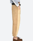 A person stands in profile wearing Sea New York’s Andrea WorkWear Pants—tan patchwork with a barrel-leg silhouette—paired with a light blue button-up and brown boat shoes, left hand in pocket against a plain white background.
