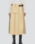 A person wearing the Sea New York Andrea Workwear Skirt, featuring a pleated midi design with a front pocket flap and leather belt, paired with a white textured long-sleeve top and black knee-high boots.