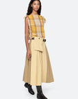 A woman in a yellow plaid sleeveless top, beige and tan Sea New York Andrea Workwear Skirt with leather belt and pouch, and black ankle boots stands against a plain light background.
