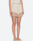 A woman wears Sea New York Ethel Solid Shorts—cream, lace with a scalloped hem and elastic waistband—styled with beige ballet flats, standing before a light gray background.