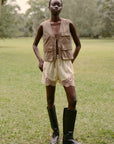 A person stands on grass outdoors, wearing the Ethel Solid Shorts by Sea New York, a tan sleeveless vest, and black knee-high boots. Trees are visible in the blurred background.