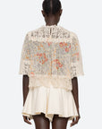A person facing away wears the Luisa Print SS Top by Sea New York, a relaxed fit sheer lace top with colorful floral embroidery and cream crochet details over a short cream skirt. The top has scalloped edges. The background is plain white.