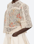 A person wears the Luisa Print SS Top by Sea New York, a cream-colored relaxed-fit lace crochet piece with floral embroidery and short sleeves, paired with cream high-waisted shorts. Their hand is in their pocket.