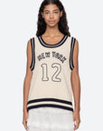 A woman with long curly hair wears the Sea New York Maren Knit Tank, a sleeveless cream cable-knit vest with black trim and "NEW YORK 12" on the front, styled over a white lace skirt against a plain light background.