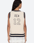 A woman with wavy dark hair faces away, wearing the Sea New York Maren Knit Tank—a sleeveless cream cable-knit vest with navy trim and "SEA 12" in navy on the back—paired with a white skirt.