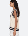 A woman with long, curly hair is shown in profile wearing the Sea New York Maren Knit Tank—a cream-colored, cable-knit vest with black trim—paired with a white lace skirt against a plain light background.