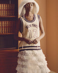 A woman stands indoors in the Maren Knit Tank by Sea New York, styled with a wide-brim hat and a striped lace skirt. Behind her, a wooden bookshelf is filled with old books.