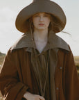 A person wears the Lois Twill Hat by Sea New York and a loose brown coat, standing outdoors in a natural, softly blurred setting and looking directly at the camera with a neutral expression.