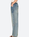 A person in Sea New York’s Claira Mended Jeans—relaxed fit with distressed patchwork and frayed hems—stands sideways, one hand in pocket, paired with open-toe heels.