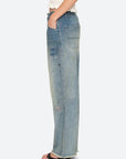 A person in Sea New York’s Claira Mended Jeans—relaxed fit with distressed patchwork and frayed hems—stands sideways, one hand in pocket, paired with open-toe heels.