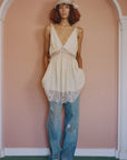 A person with curly hair wears Claira Mended Jeans by Sea New York, a lacy cream dress, sandals, and a decorative headpiece, posing against a pink wall framed by a white arch.