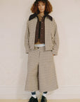 Wearing the Savannah Plaid Culotte by Sea New York, a person with curly hair pairs it with a loose checked jacket, brown shirt, visible boxer-style waistband, striped socks, and black shoes while standing against a pale wall.