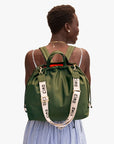 A person with short hair, in a sleeveless light blue and white striped dress, carries Clare Vivier’s Sac Tout Backpack in olive recycled nylon with cream “ciao” straps, gold hardware, and black cords.