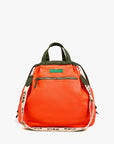The Sac Tout Backpack by Clare Vivier is a large orange recycled nylon bag with green handles, a front zip pocket, and a detachable beige "Ciao" shoulder strap in black print, shown on a plain white background.