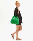 A woman with curly blonde hair in a bun wears a black blazer, black shorts, and black sandals, standing sideways with the Clare Vivier Sac Tout—a large green nylon drawstring bag—over her shoulder against a white background.