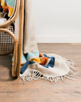 The Sackcloth & Ashes/Camp Coast blanket by Sackcloth & Ashes, cream-colored with colorful geometric patterns and fringe, drapes over a wicker chair onto a wooden floor against a plain white wall.
