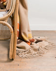The Sackcloth & Ashes/Desert Camp blanket by Sackcloth & Ashes, with its colorful fringe, drapes over a woven wooden chair on a light wood floor—an inviting accent for any room.