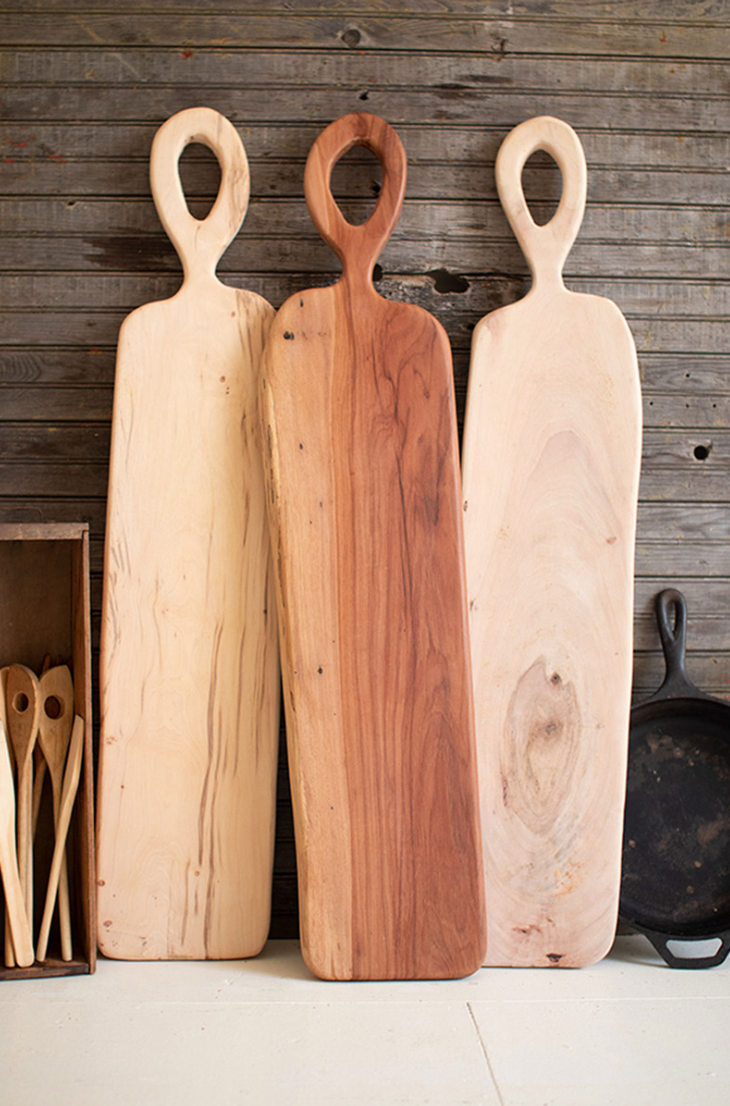Three tall Kalalou, Inc Pecan Charcuterie Boards with elongated handles lean against a rustic wooden wall in a Scottsdale, Arizona bungalow, near wooden spoons and a black cast iron skillet.