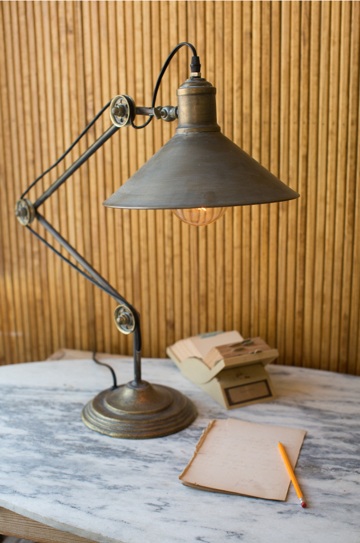 An antique-style Metal Table Lamp with an extended arm illuminates a notebook and a pencil on a marble table, with a stack of books nearby, against the background of a wooden wall in a Scottsdale Arizona bungalow.