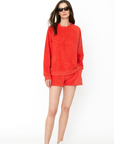 A woman in a vibrant red sweater and Kule's Terry Venus shorts set, wearing white sneakers and sunglasses, stands confidently against a white background.