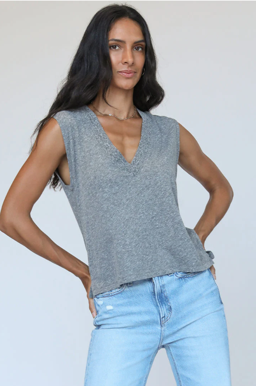 A woman with long dark hair wearing a Margot Cotton Sleeveless V-Neck Tee by perfectwhitetee and blue jeans stands confidently with hands on hips in front of a bungalow.
