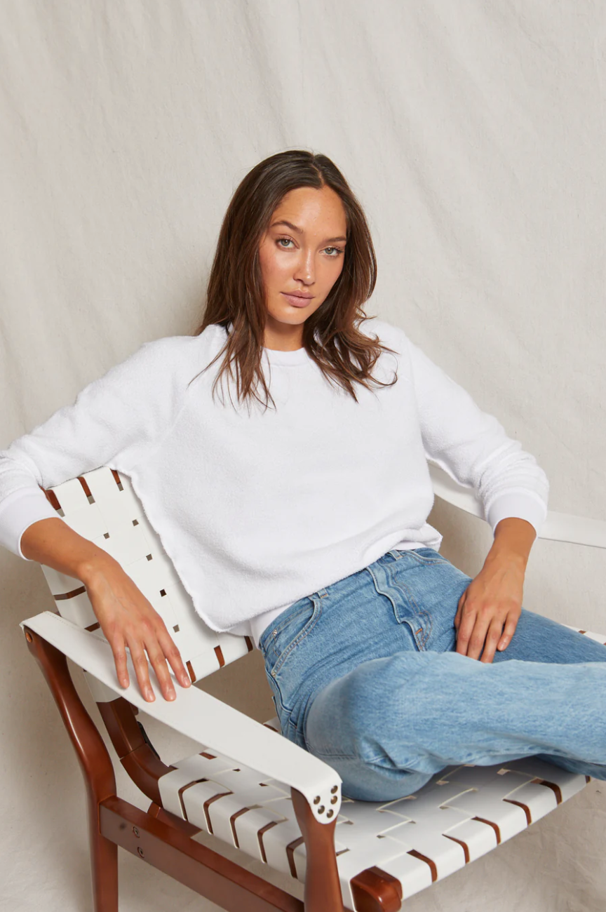 A woman sits relaxed in a white chair in her bungalow, wearing a Perfectwhitetee Ziggy Reverse Fleece LS Shrunken Crewneck Sweatshirt and blue jeans. She gazes at the camera with a composed expression, her long brown hair loosely framing her face.