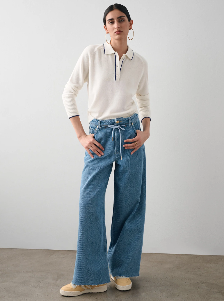 A woman stands confidently in Scottsdale, Arizona, wearing a White + Warren Cashmere Ribbed Trim Polo and wide-legged blue jeans, accessorized with large hoop earrings, against a neutral background.