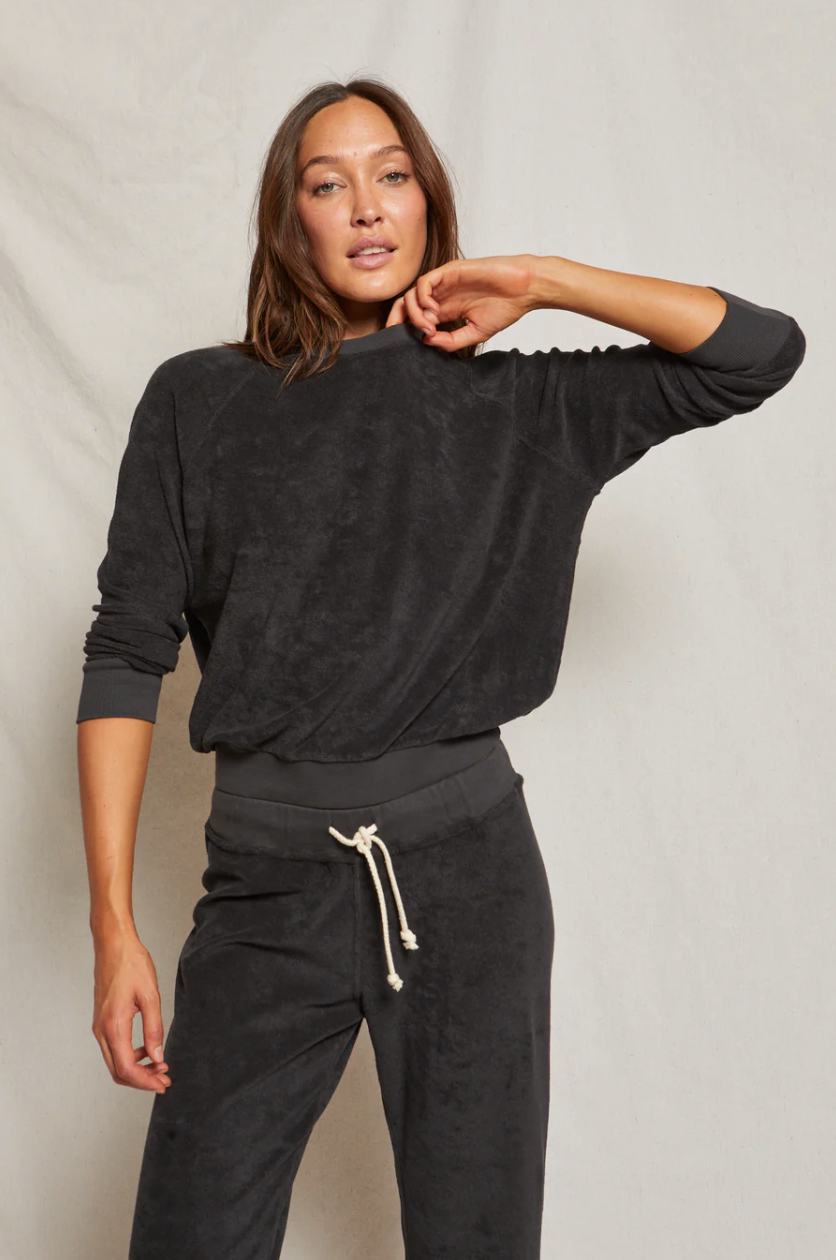A woman in a Perfectwhitetee Saylor Loop Terry Sweatshirt and matching sweatpants posing against a light beige background in her bungalow, her hand resting on her neck.