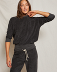A woman in a Perfectwhitetee Saylor Loop Terry Sweatshirt and matching sweatpants posing against a light beige background in her bungalow, her hand resting on her neck.