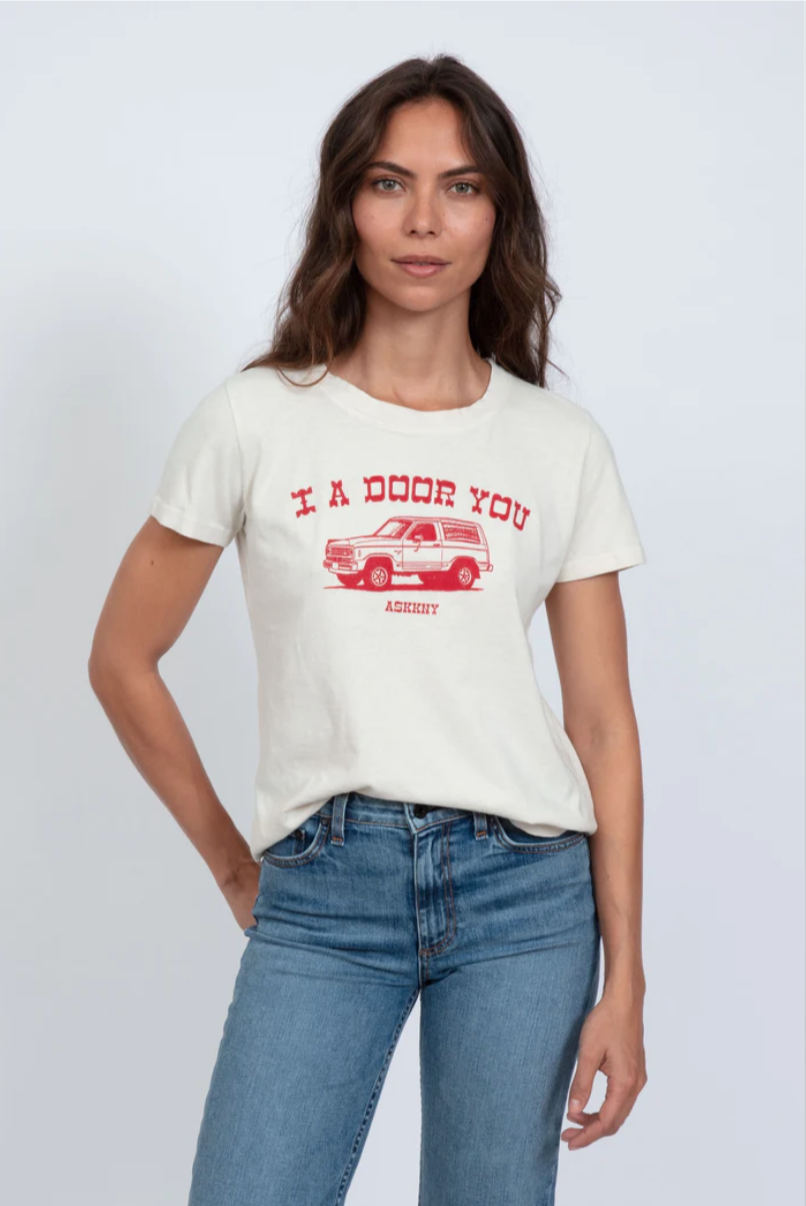 A woman with brown hair stands against a white background, wearing a white ASKK printed classic tee with a red bungalow and the text "I A-DOOR YOU" printed on it, paired with blue jeans.
