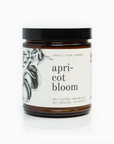A Faire soy candle in a clear glass jar with a label that reads "Apricot Bloom" and includes illustrations of fruit and flowers. The label mentions cotton, natural oils, and a 50-hour burn time.