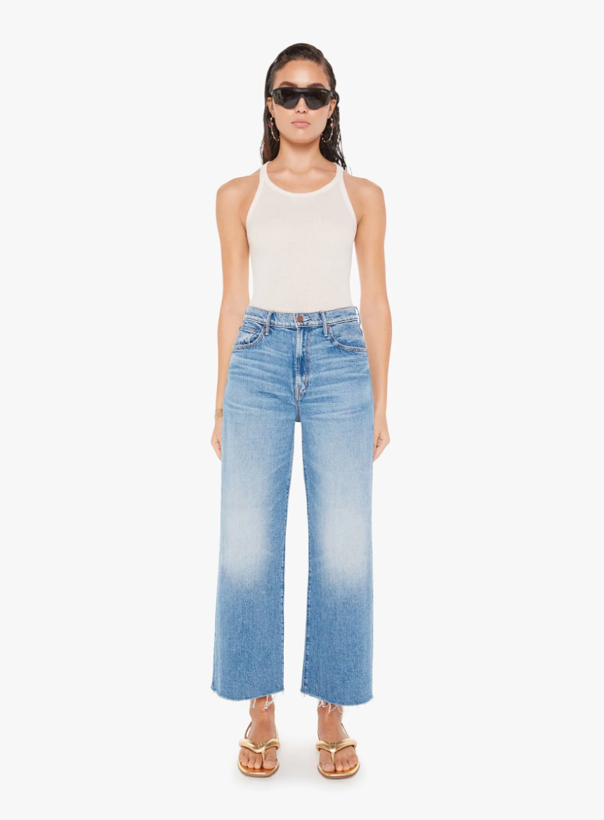 A woman stands confidently wearing Mother's The Maven Ankle Fray jeans, a white tank top, high-waisted mid-blue wash jeans, and strappy sandals. She poses against a plain white background.
