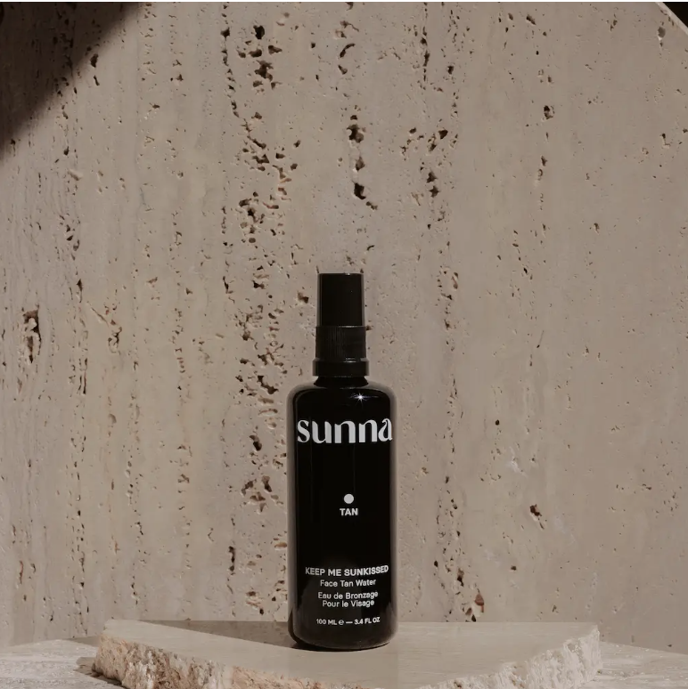 A black spray bottle labeled &quot;Faire Face Tan Water&quot; stands on a stone slab against a textured beige concrete wall in Scottsdale, Arizona. The label reads &quot;Keep me sunkissed, Face &amp; Body Water, 100 ml e 3.38 fl oz.