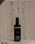 A black spray bottle labeled "Faire Face Tan Water" stands on a stone slab against a textured beige concrete wall in Scottsdale, Arizona. The label reads "Keep me sunkissed, Face & Body Water, 100 ml e 3.38 fl oz.