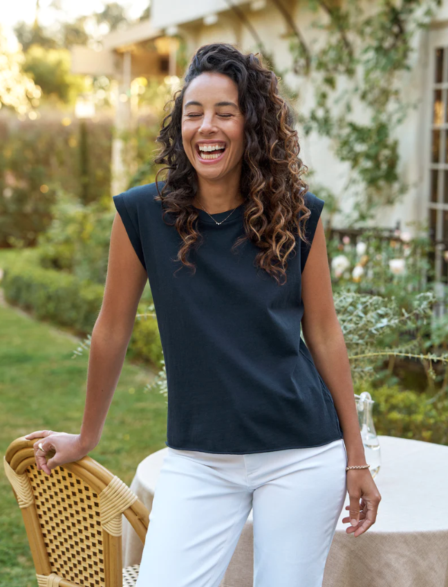 A woman with curly hair, wearing a AIDEN Vintage Muscle Tee HERITAGE JERSEY by Frank &amp; Eileen and white pants, laughs joyfully while standing in the garden of a Scottsdale, Arizona bungalow next to a wicker chair.