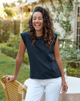 A woman with curly hair, wearing a AIDEN Vintage Muscle Tee HERITAGE JERSEY by Frank & Eileen and white pants, laughs joyfully while standing in the garden of a Scottsdale, Arizona bungalow next to a wicker chair.