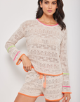 A woman models a beige crochet Beach Please Popover sweater and matching shorts with neon pink and orange trim, embodying Arizona-inspired style. She stands confidently against a plain background, looking directly at the camera.