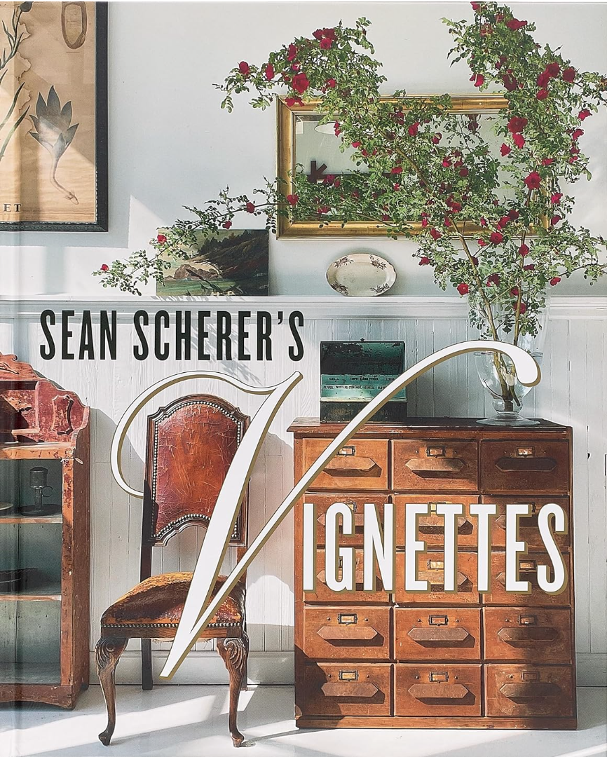 Book cover for "Sean Scherer's Vignettes" featuring a rustic and eclectic interior from Hachette Book Group, with a wooden chair, a chest of small drawers, botanical prints, and a floral arrangement on a white wainscot backdrop. Elegant, large text overlays the central part of the cover.