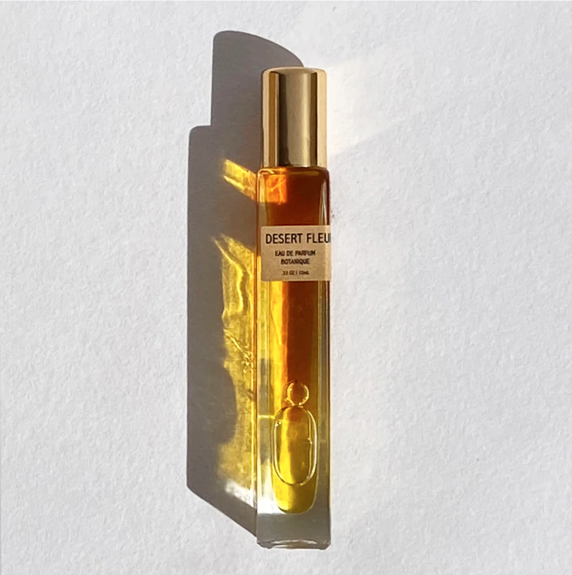 A bottle of "Desert Fleur Parfum" by Faire against a white background, photographed in a Scottsdale Arizona bungalow, with sunlight casting sharp shadows to the left. The perfume liquid is amber-colored.