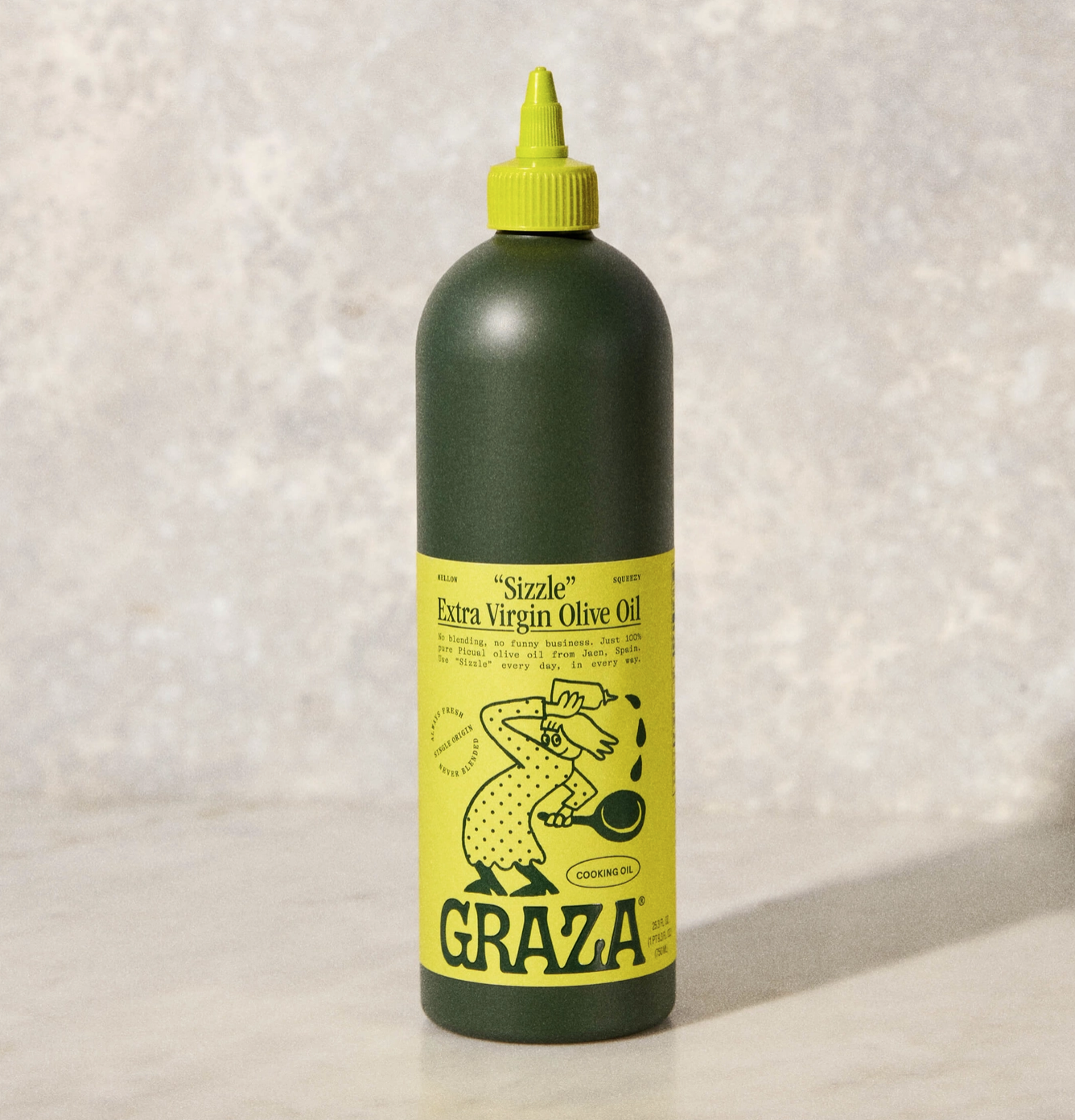 A bottle of Faire &quot;Sizzle&quot; extra virgin olive oil with a playful label featuring a yellow illustration of a chicken cooking at a bungalow, set against a textured gray background.