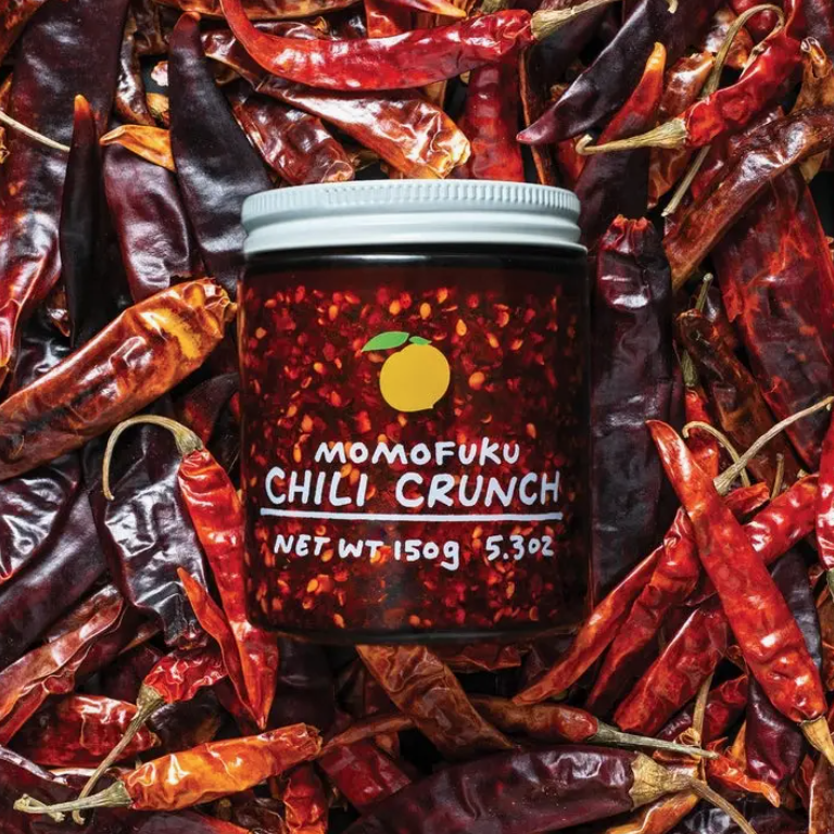 A jar of Faire Original Chili Crunch is centered among scattered dried red chili peppers on the porch of a Scottsdale, Arizona, bungalow. The jar features a label with a peach logo above the product name.