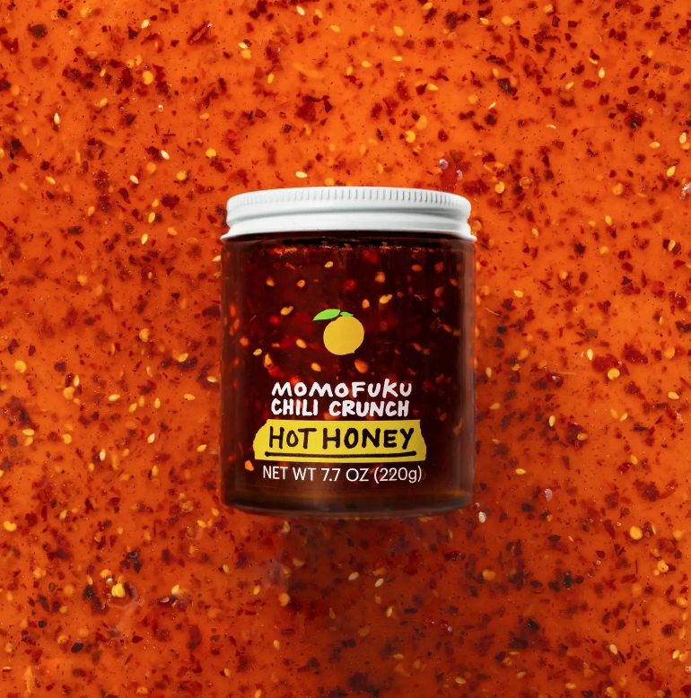 A jar of Faire Chili Crunch Hot Honey sits centered on a textured orange background speckled with red chili flakes, reminiscent of a vibrant sunset in Scottsdale, Arizona.