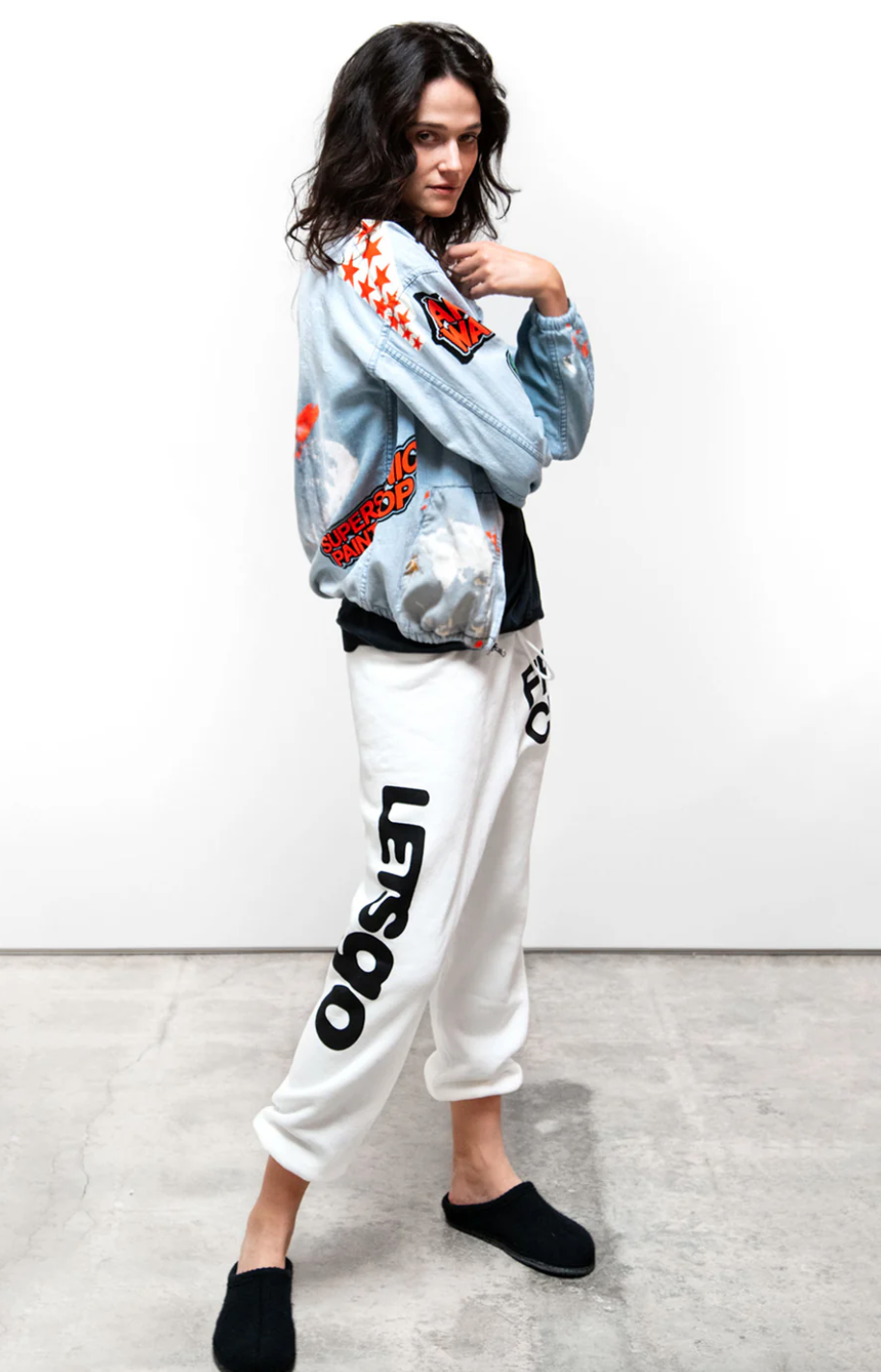 A woman stands confidently wearing a stylish white and red graphic jacket over a T-shirt, paired with the CIRCA&#39;99 OG LETSGO OLDSCHOOL POLYBLEND/FLUFF sweat sweatpants marked &quot;LET&#39;S GO&quot; in black letters, and black slip-on shoes, against a plain background.