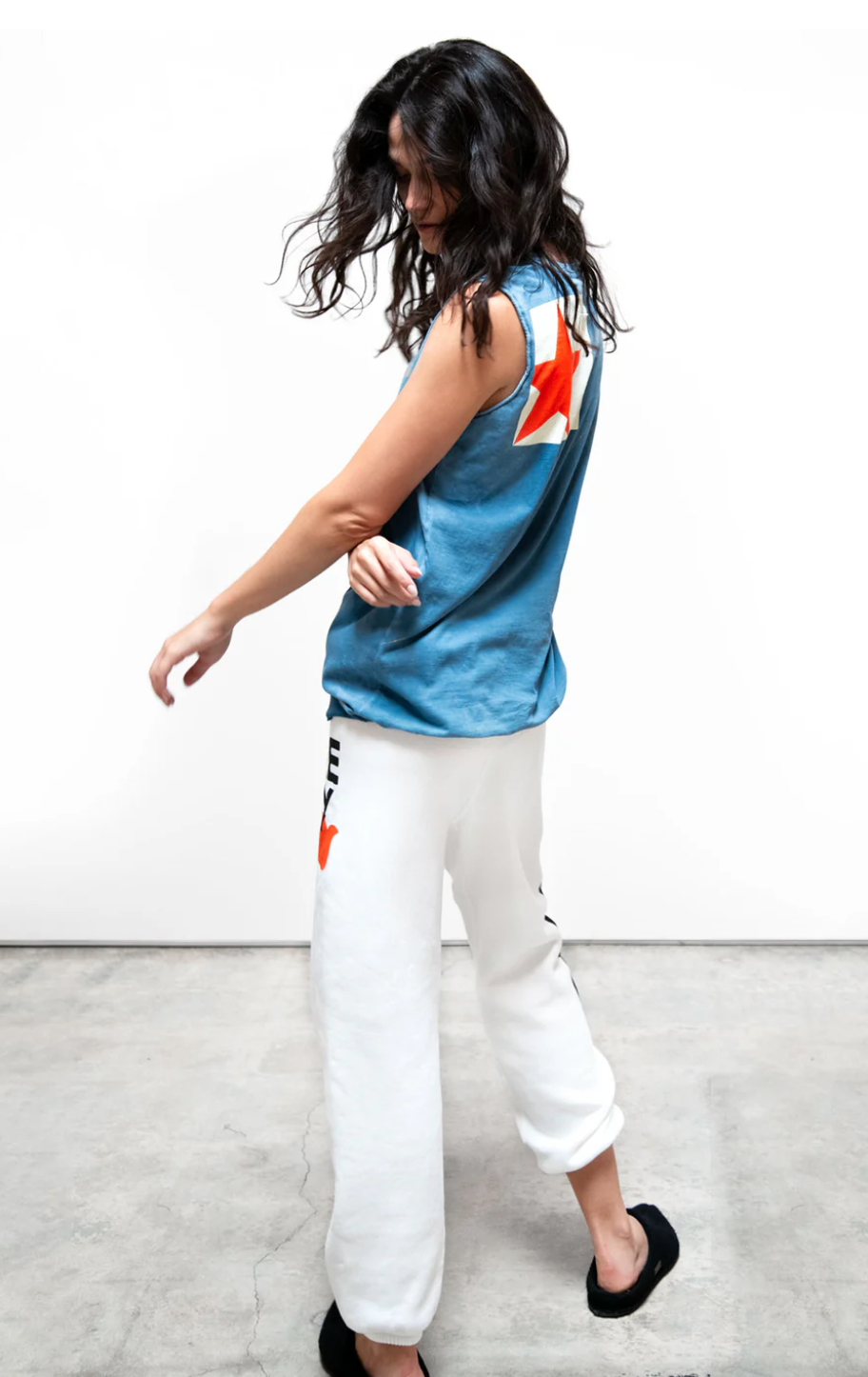 A woman with long dark hair dances joyfully, wearing a Free City (sparrow, LLC) GOLDEN SIGN/STAR BIGGY tank/lux rib with a lightning bolt design and white sweatpants, against a plain white background in Arizona style.