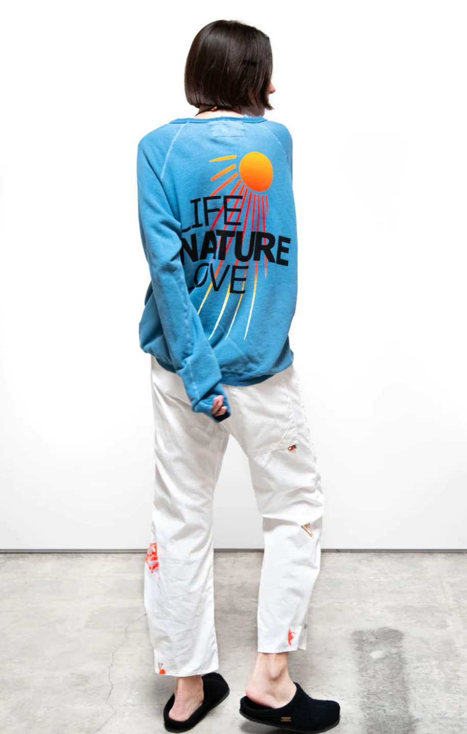 A person stands with their back towards the camera, wearing a LNLSUN SUPERYUMM BIGGIE raglan sweatshirt from Free City (sparrow, LLC) with the text "LIFE, NATURE, LOVE" and a graphic of a sun, paired with white, distressed jeans and black slippers in Arizona style.